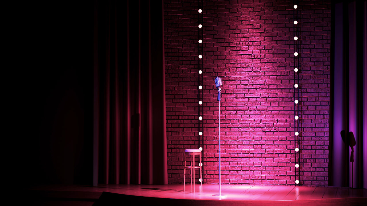 A microphone and stool on stage under a purple spotlight with a curtain to the left
