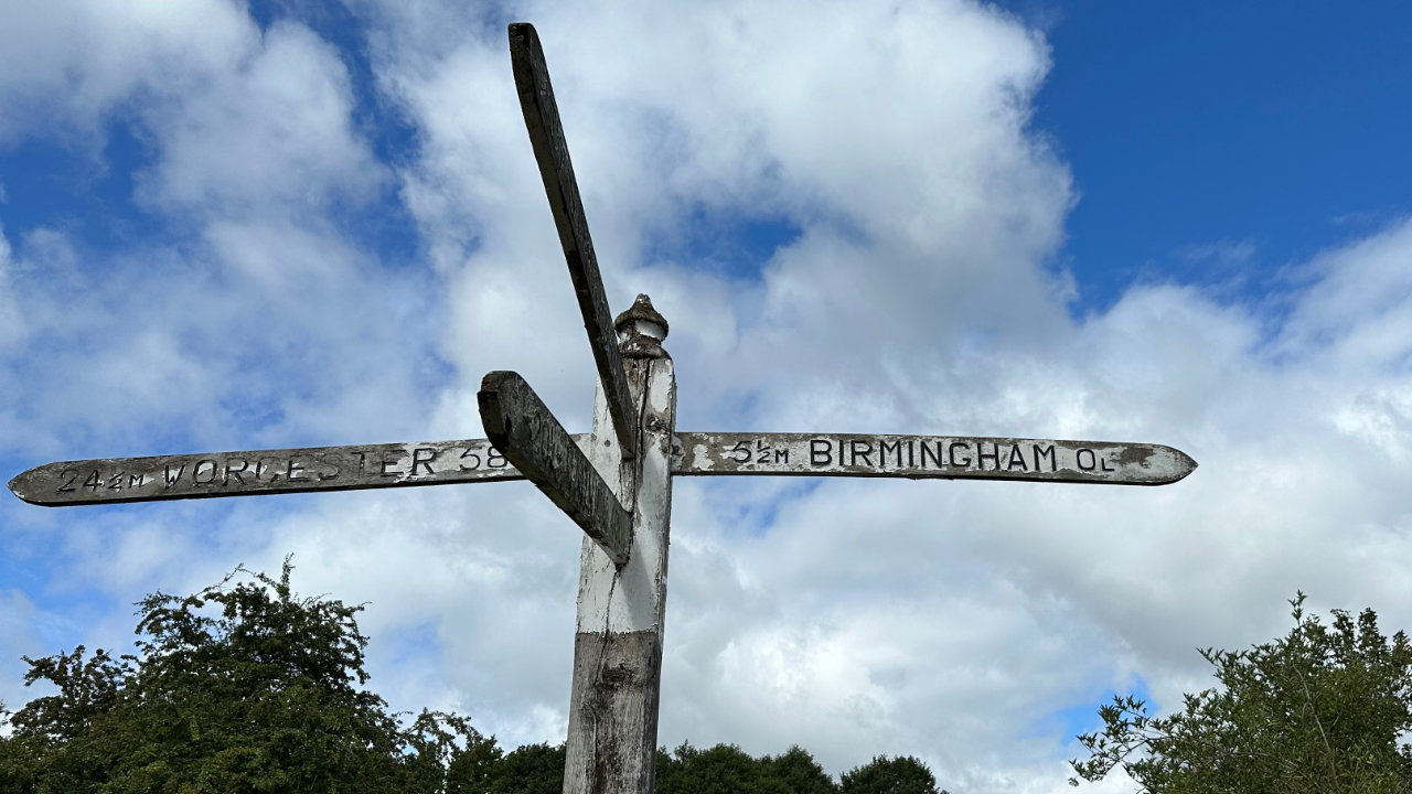 A blue sky with green trees in the background, and in the foreground a battered old signpost pointing to Birmingham on the right and Worcester on the left