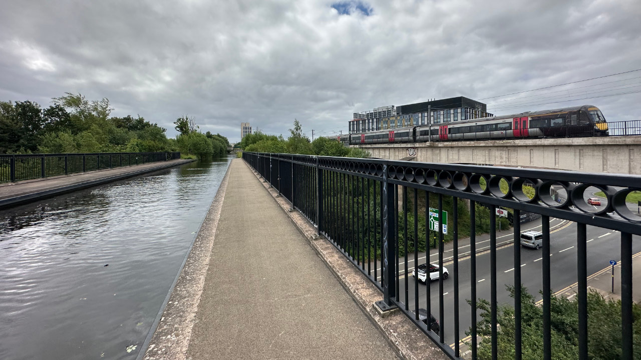 A photo taken from the point of view of the tow path of a canal over an aqueduct, with a dual carriageway passing underneath and a couple of cars travelling to the right. A railway bridge is also on the right with a train just approaching towards the camera.