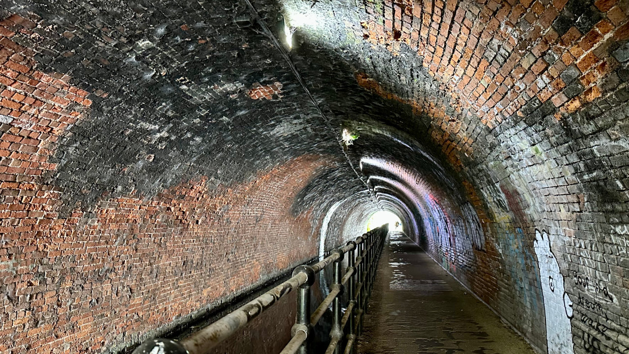 A perspective photo inside a canal tunnel, with red brick arch over the water, a black rail and black tow path to the right hand side, all leading towards the light at the end of the tunnel in the distance