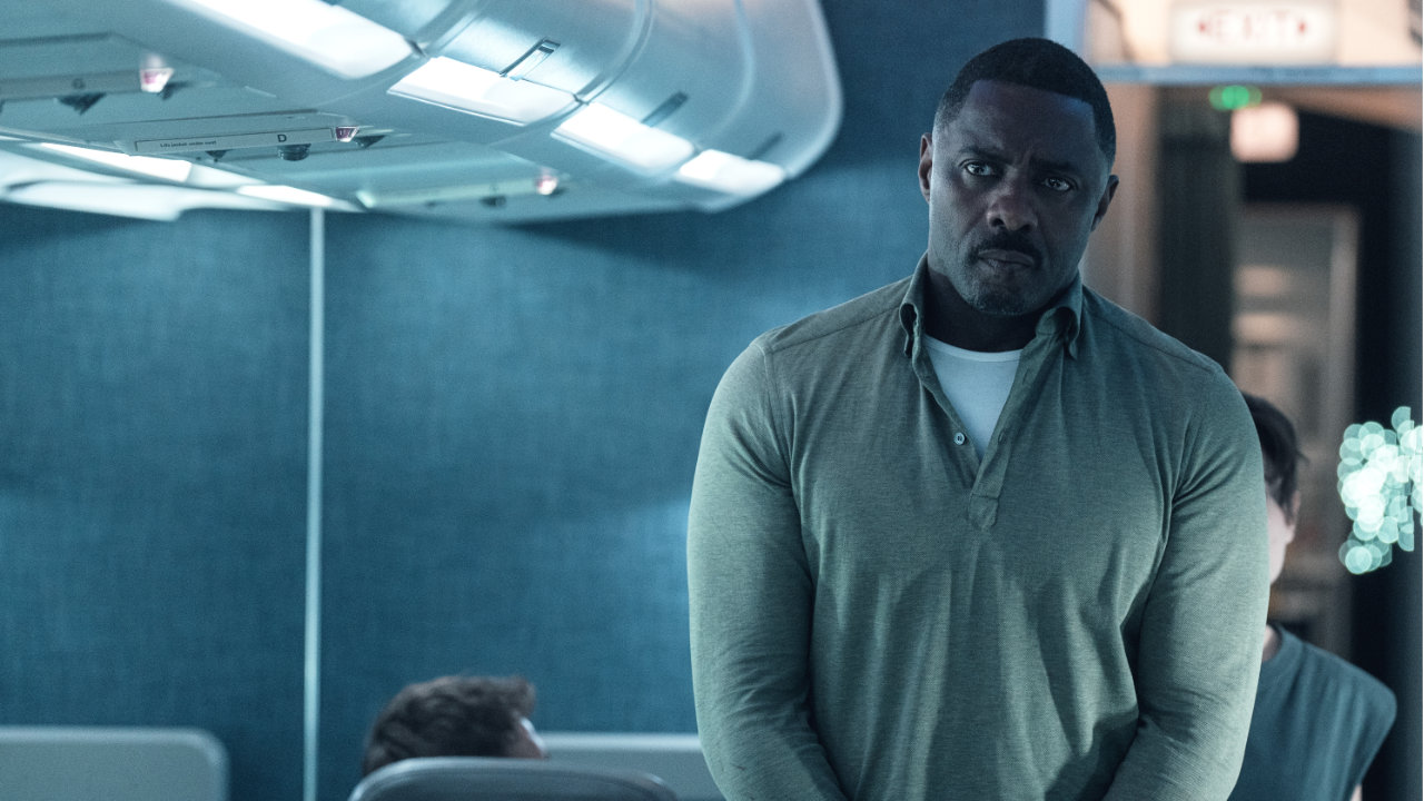 A screenshot from the Apple TV series Hijack, featuring Idris Alba in a grey jumper standing in the aisle of a plane
