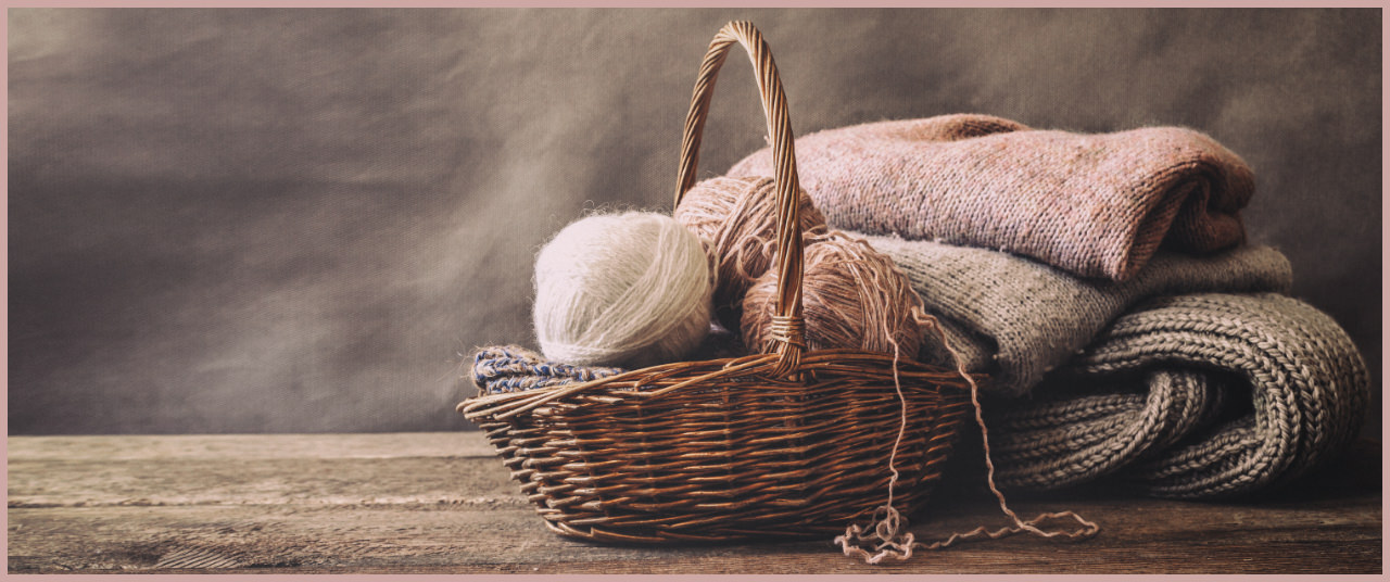A wicker basket with balls of wool next to a pile of woollen jumpers