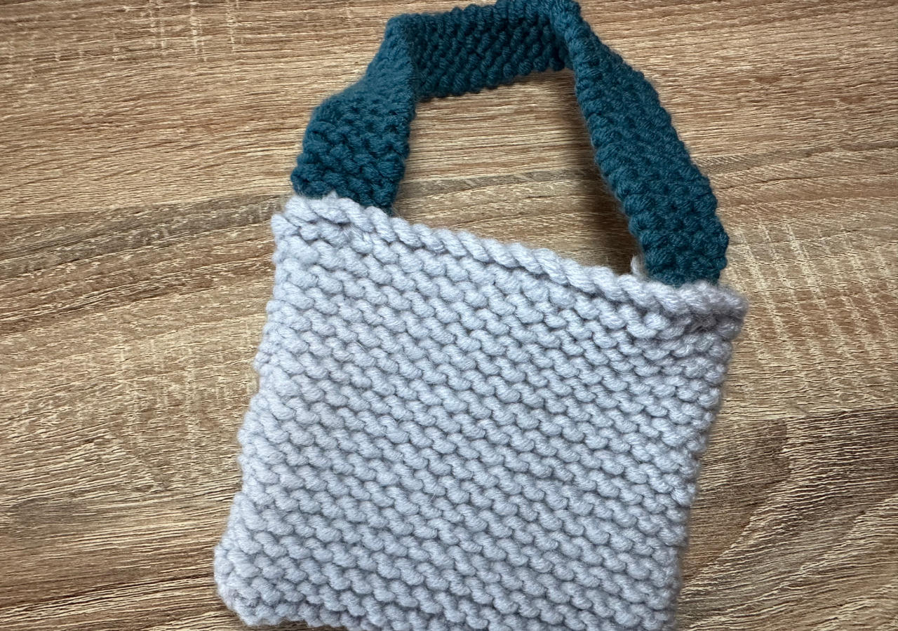 A photograph of a small knitted bag in grey with a green handle
