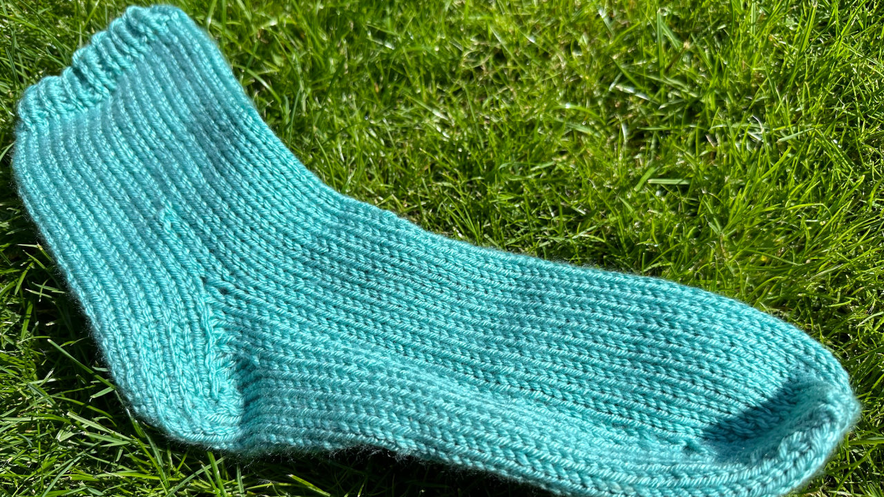 One of the knitted socks laid flat against the grass, showing the knitted heel and toe detail