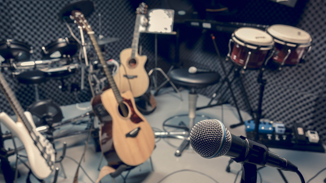 A music studio with various instruments in the background - three guitars and two sets of drums - with a microphone in focus at the front