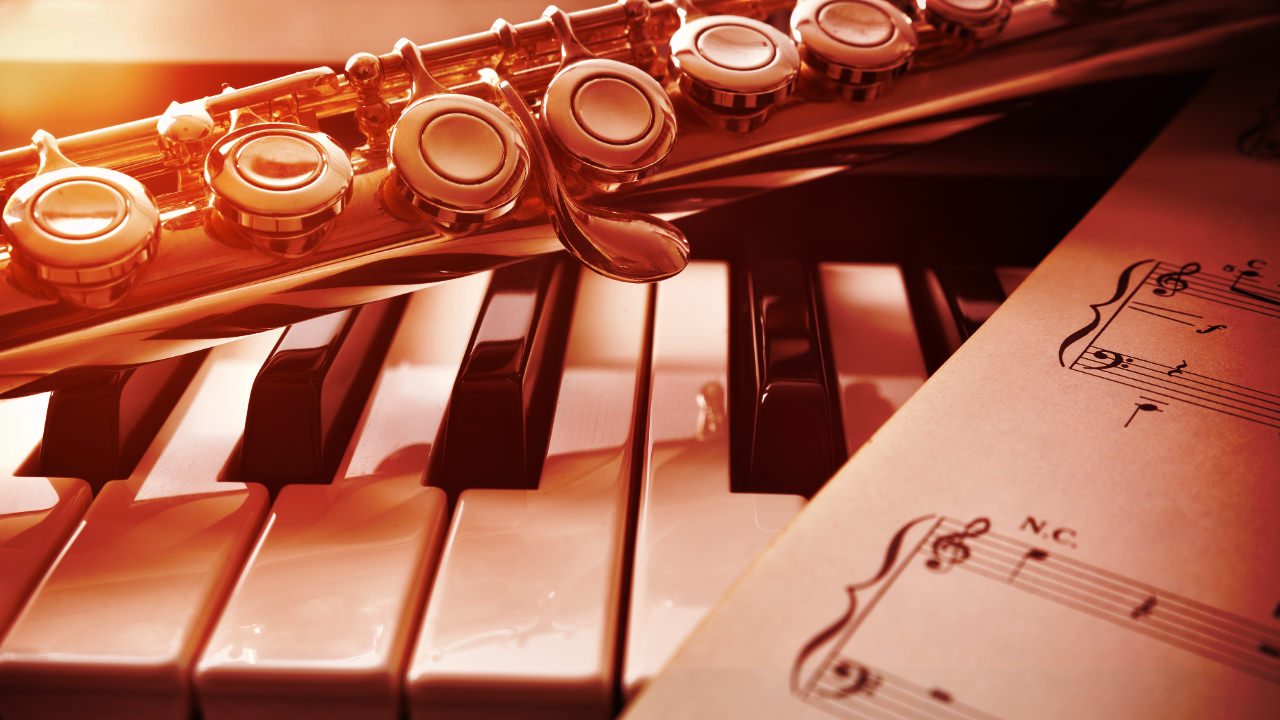 A close up of piano keys, with a flute lying on top and some sheet music to one side