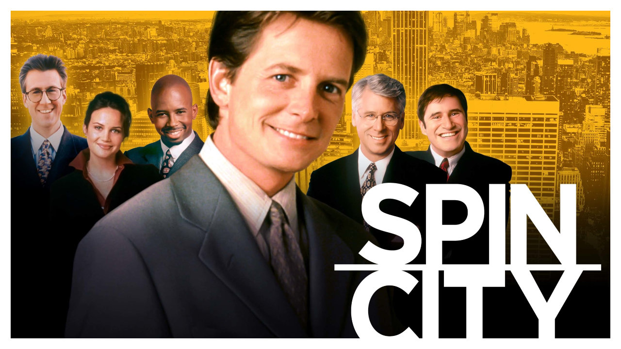 A marketing still from Spin City featuring Michael J Fox front and centre with several supporting characters behind him, the whole thing bordered in white with Spin City in white text
