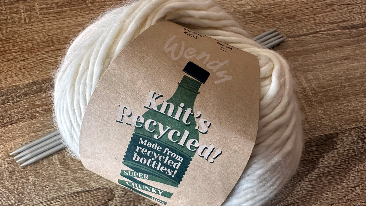 A ball of Wendy&rsquo;s recycled yarn in cream, the label saying &lsquo;Knit&rsquo;s Recycled! Made from recycled bottles! Super Chunky&rsquo; and behind it a set of four double pointed needles with the ends sticking out each side of the yarn