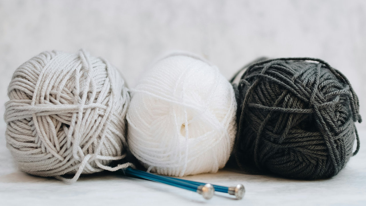 Three balls of wool in a row, grey, white and dark grey from left to right, with two blue knitting needles sitting in front of them