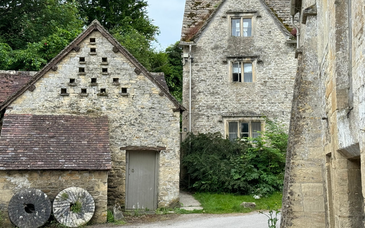 A view of the end of a country lane with several old buildings in stone and two mill wheels propped up against a wall