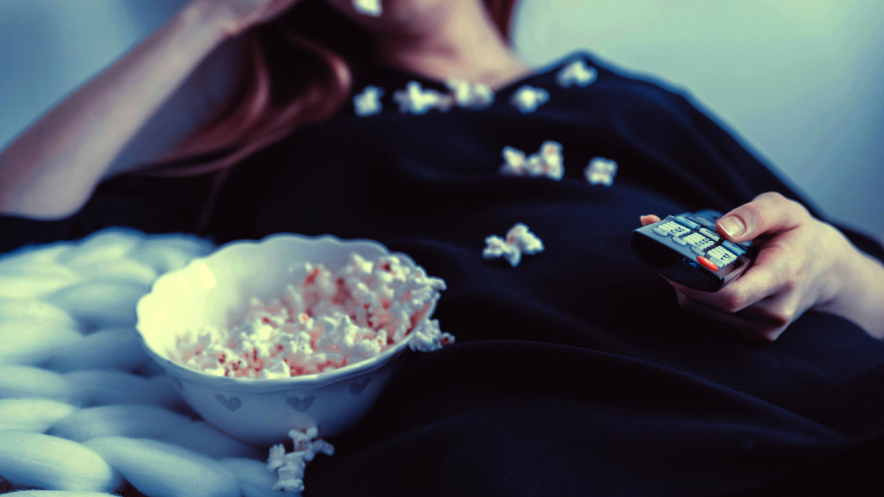 A photograph of a woman leaning back on a sofa or bed with a remote control in one hand and a bowl of popcorn next to her, kernals of popcorn scattered on her front