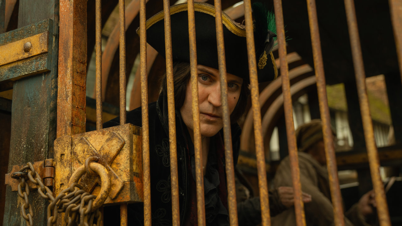 A marketing still image from the Apple TV show The Completely Made Up Adventures of Dick Turpin, featuring Noel Fielding as the title character, sitting in a carriage behind bars