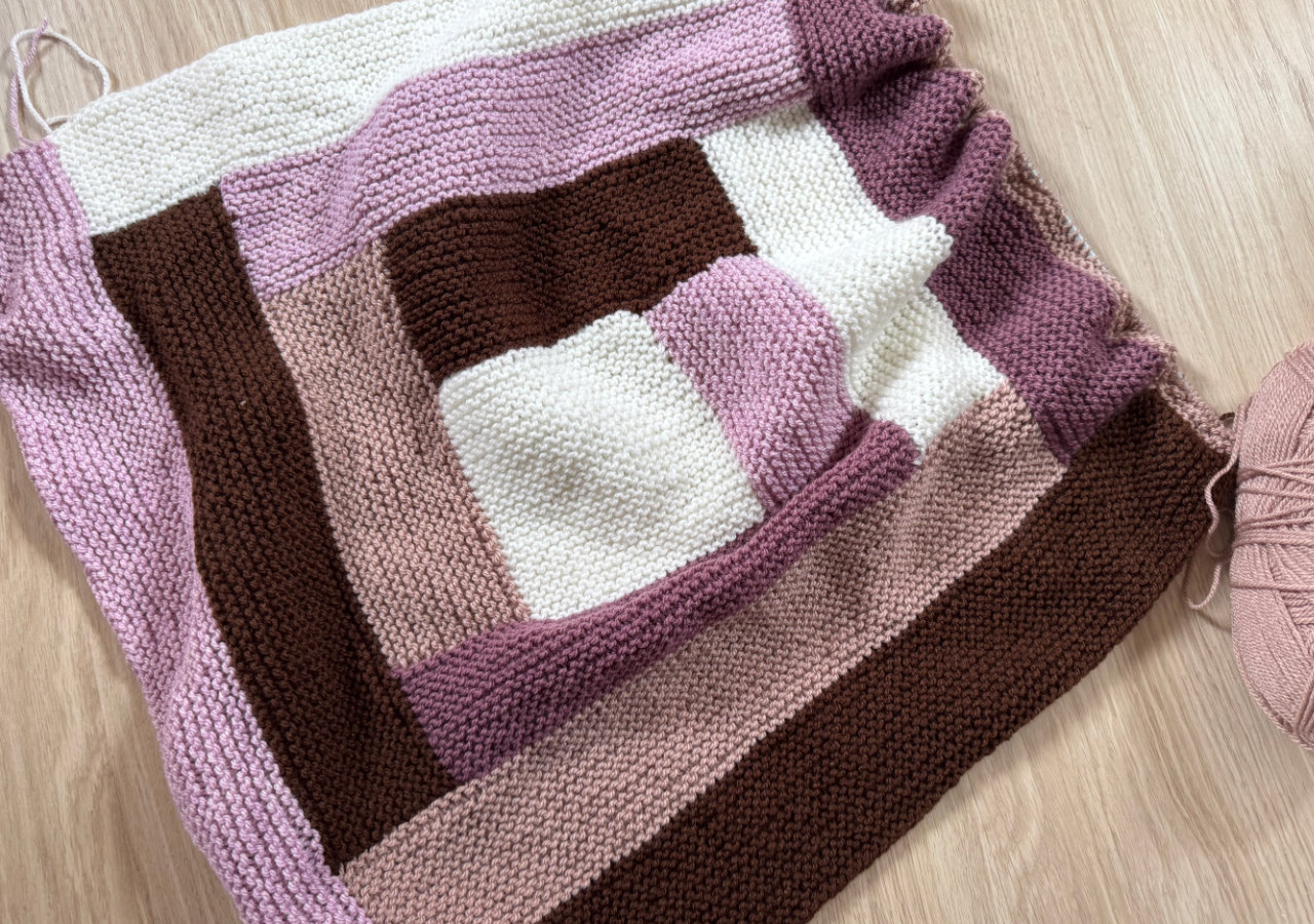 An in-progress knitted blanket featuring a white square in the middle and then stripes of brown, beige, pink and purple building up around it