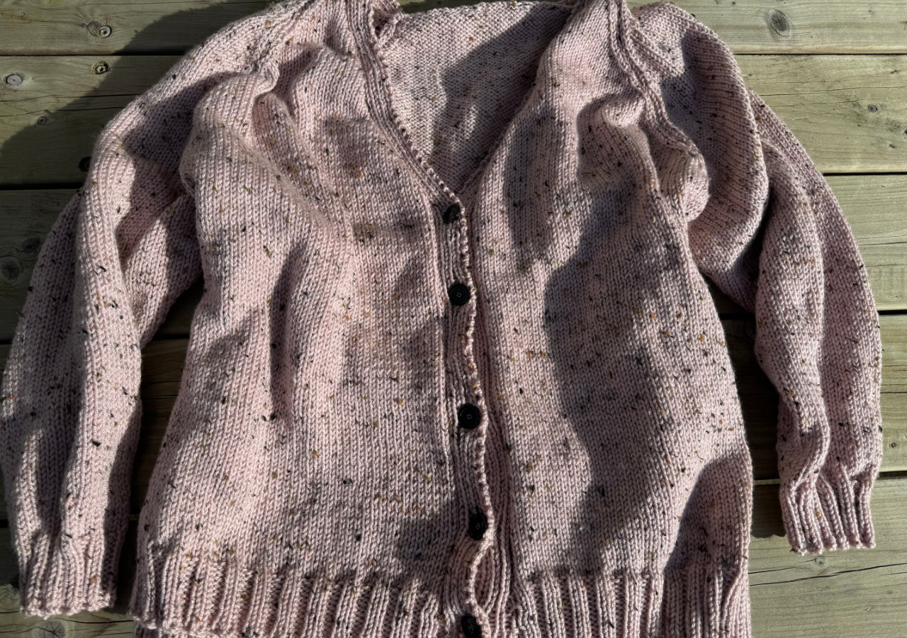 A photograph of a knitted pink cardigan with black buttons on a garden bench table