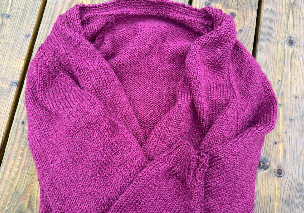 A plum knitted cardigan sitting on a wooden surface with the arms folded across the front