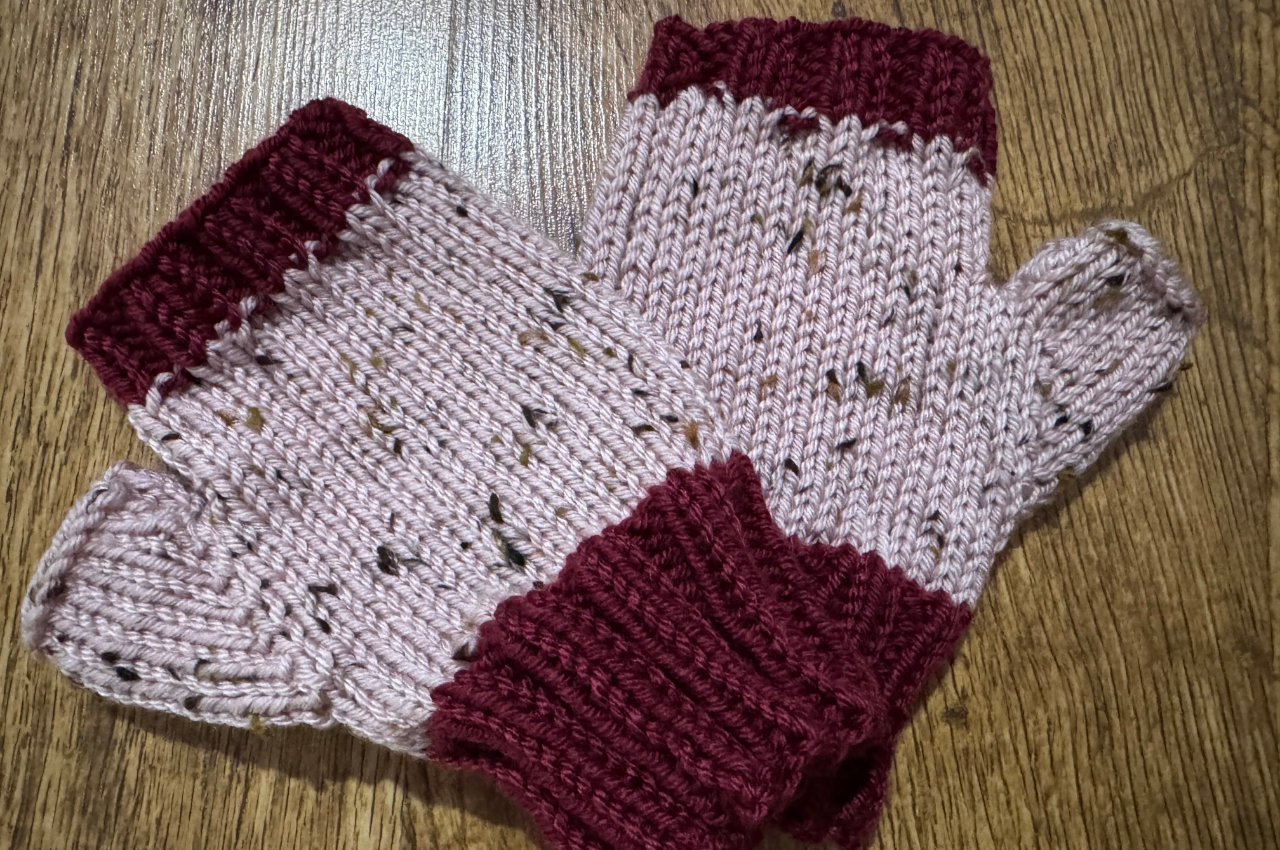 a pair of knitted fingerless mittens on a hardwood floor, the ribbing in dark red, the main body of the glove in pink tweed