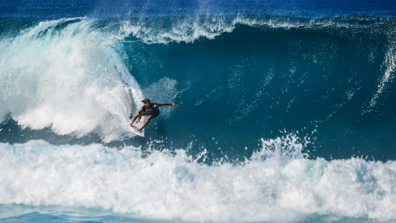 A surfer glides along a huge wave from left to right with the surf and spray following them to the left