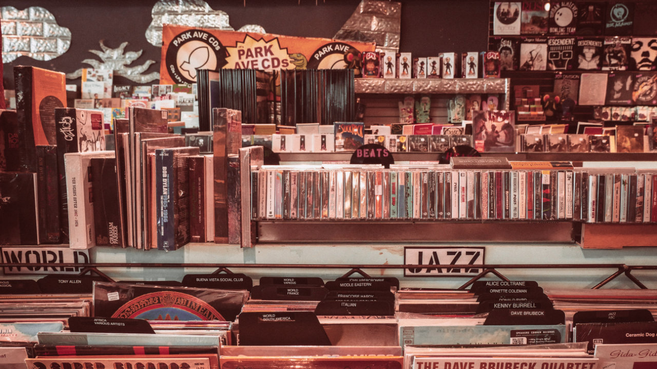 A photograph tinted sepia and pink that shows a crammed section of a record store, with tapes, records and CDs cluttered around
