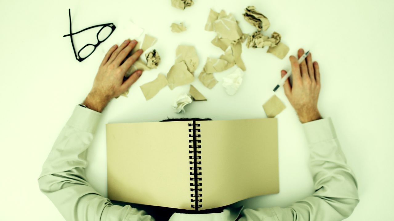 A top down photograph of an office worker with their head slumped forward on the desk, covered by an open notebook