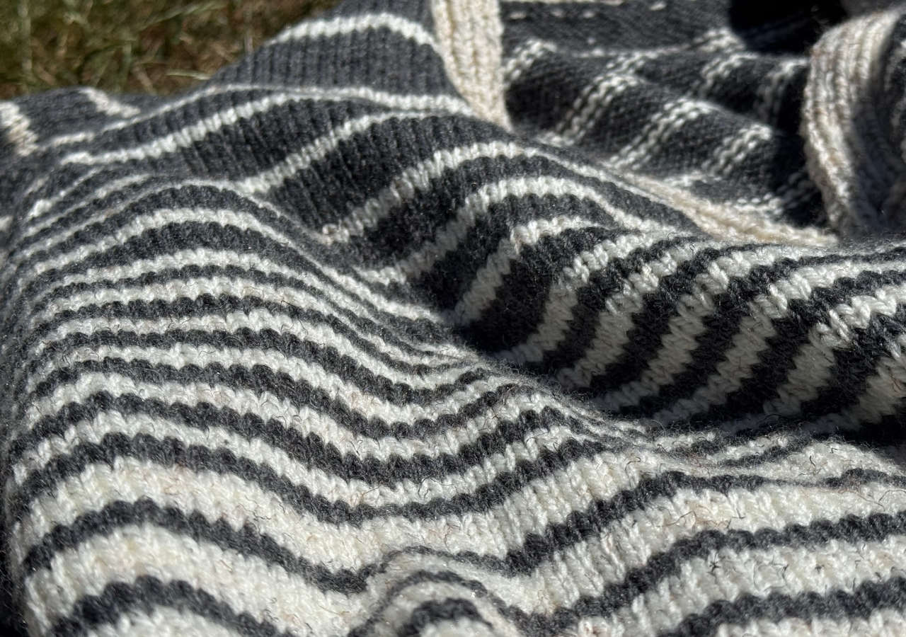A hand knitted cardigan, white and grey striped with black buttons, lying on the grass