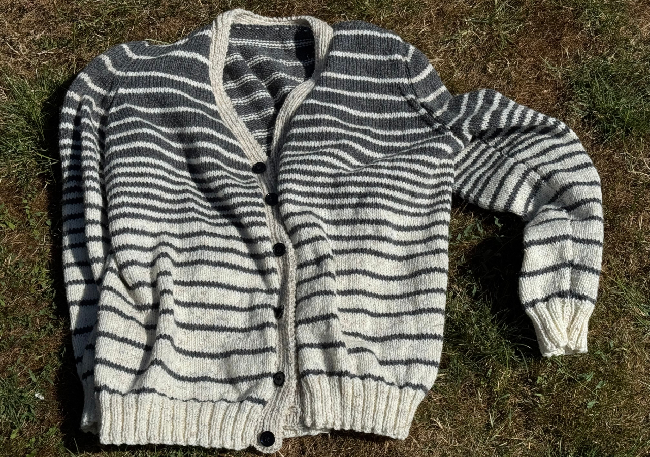 A hand knitted cardigan, white and grey striped with black buttons, lying on the grass