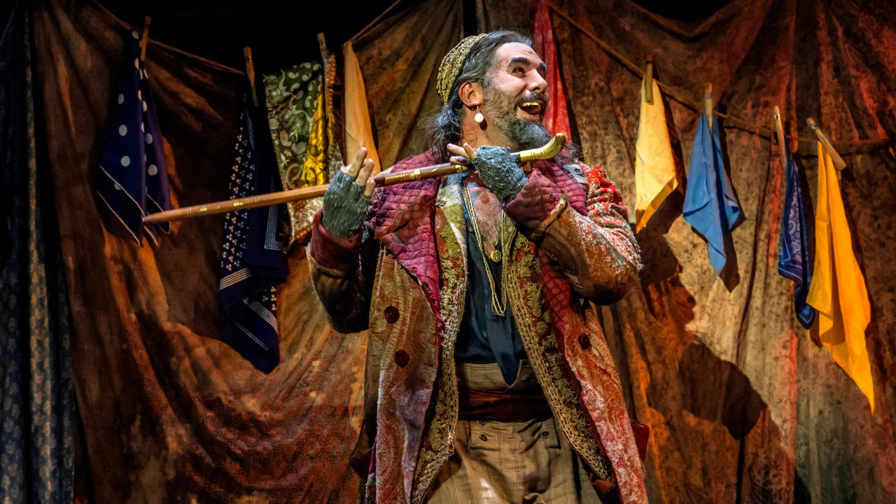 A marketing image from the London West End production of Oliver! featuring Simon Lipkin as Fagin, dressed in colourful rags and playing a pipe
