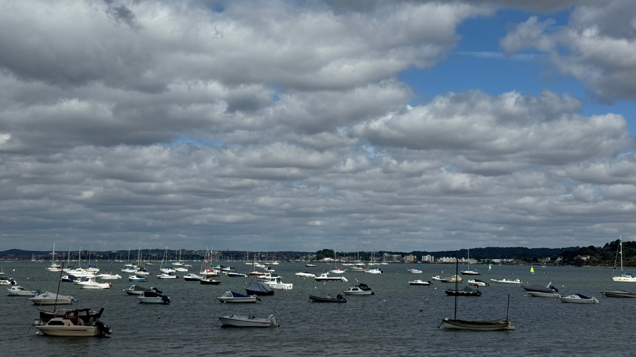 A photo of the coastline at Swanage, with lots of sailing boats dotted on the water, and fluffy rolling clouds in a large blue sky