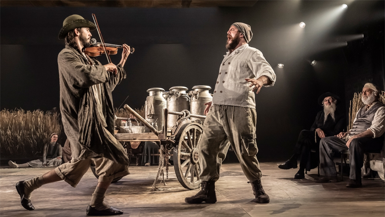 A promotional image for the UK tour of the musical Fiddler on the Roof, featuring Tevye by his milk cart, looking at the fiddler playing his violin