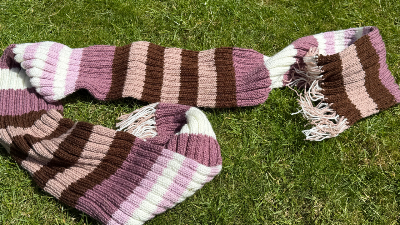 A knitted scarf, striped with purple, brown, cream and pink colours, lying on the grass